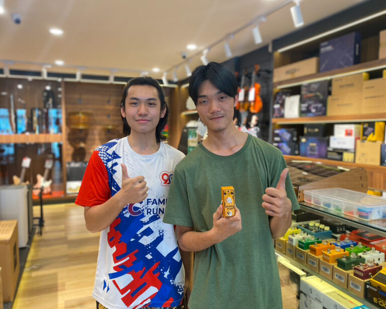 Two young men in a music store posing for a photo, giving thumbs up; one holds a small yellow boxed item.
