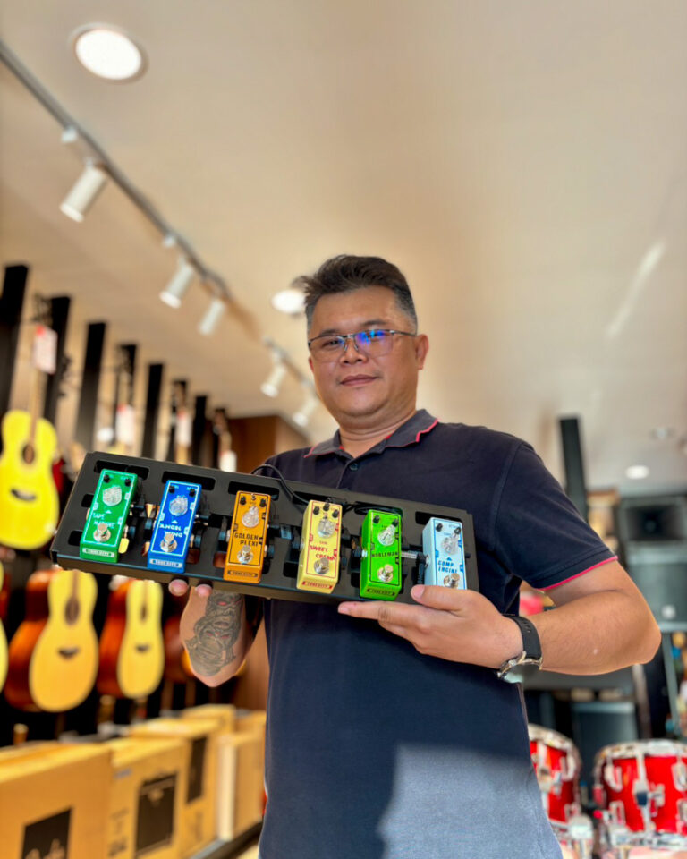 Man in a dark polo holds a black pedalboard with seven colorful guitar effect pedals in a music store.