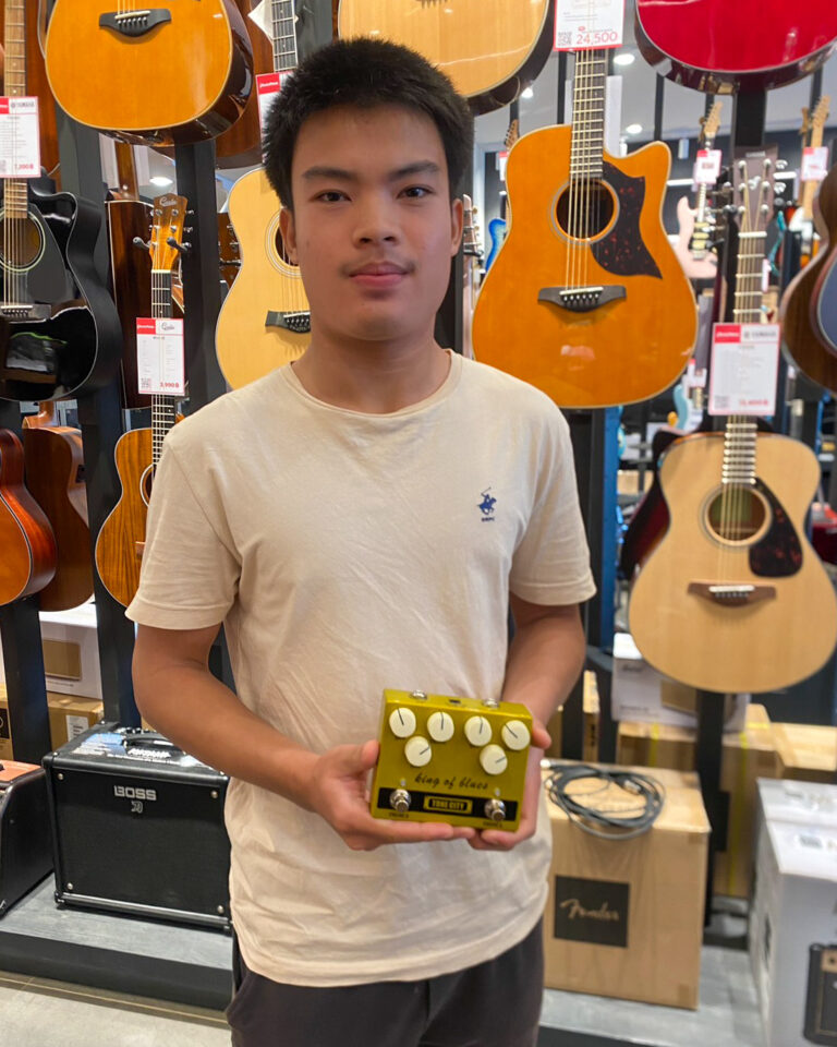 Young man in a guitar store holding a yellow guitar effects pedal, with acoustic guitars hanging on display behind him.