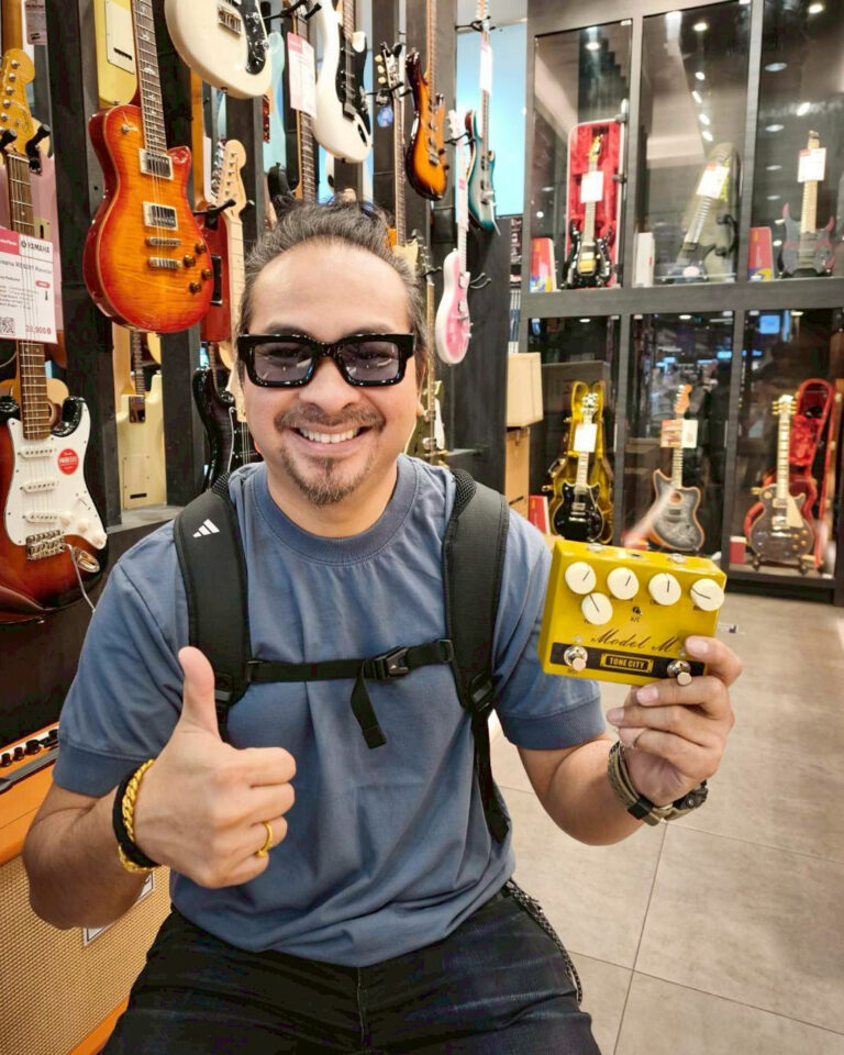 Man in a guitar shop giving a thumbs up while holding a yellow Model M guitar effects pedal.