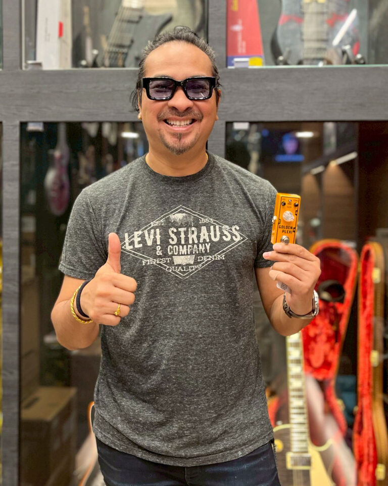 Smiling man in sunglasses gives a thumbs up while holding an orange guitar effects pedal in a music store nearby guitars display in the background.