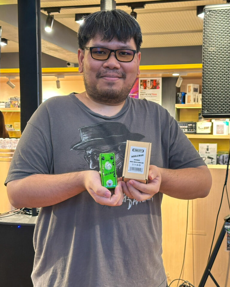 Smiling man with glasses in a music store holds a green Nobelman guitar pedal and a small brown box.