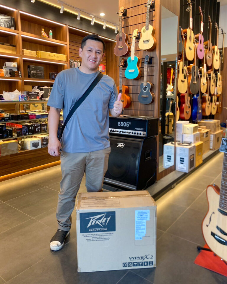 Smiling man in a blue shirt gives a thumbs-up inside a guitar shop, surrounded by wall-mounted guitars and audio gear; a large Peavey box sits on the floor.