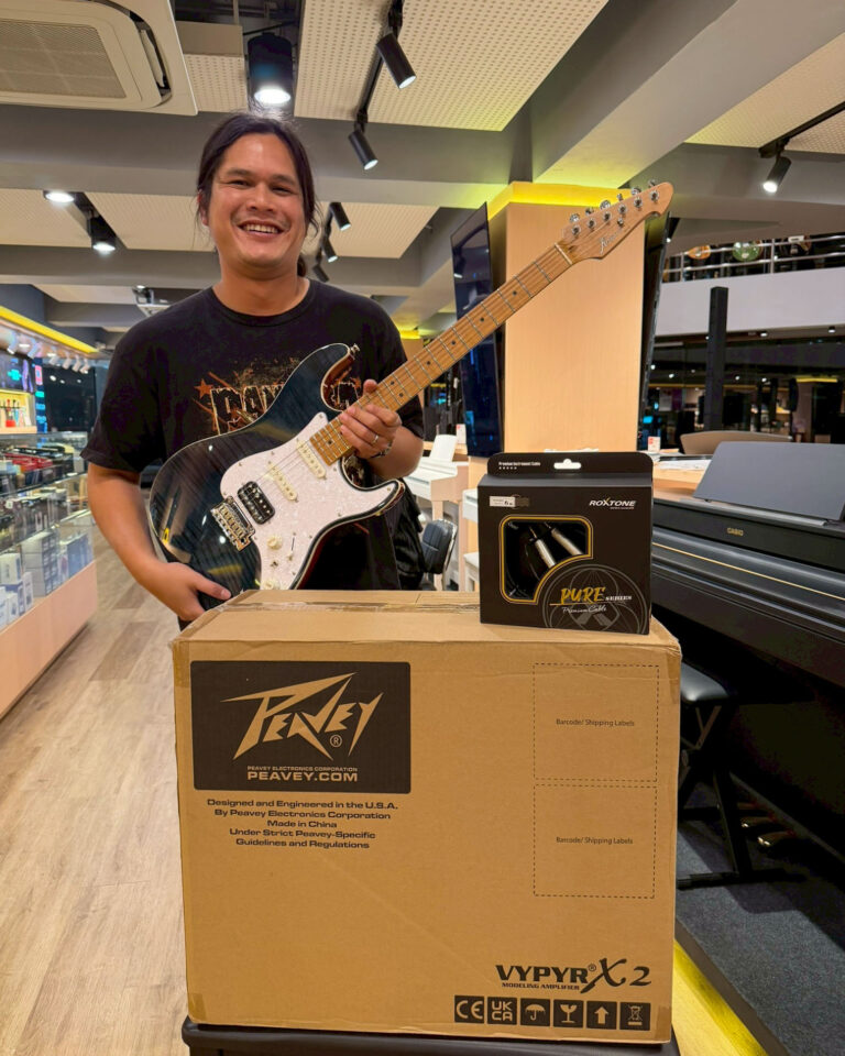 Smiling man in a store holding a black electric guitar next to large guitar amplifier and packaging boxes on a checkout counter.