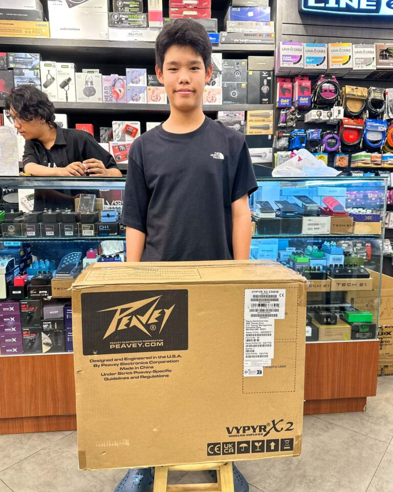 Teenager in a black t-shirt stands behind a glass display counter in an electronics store with shelves of audio gear behind him and a large Peavey box in front.