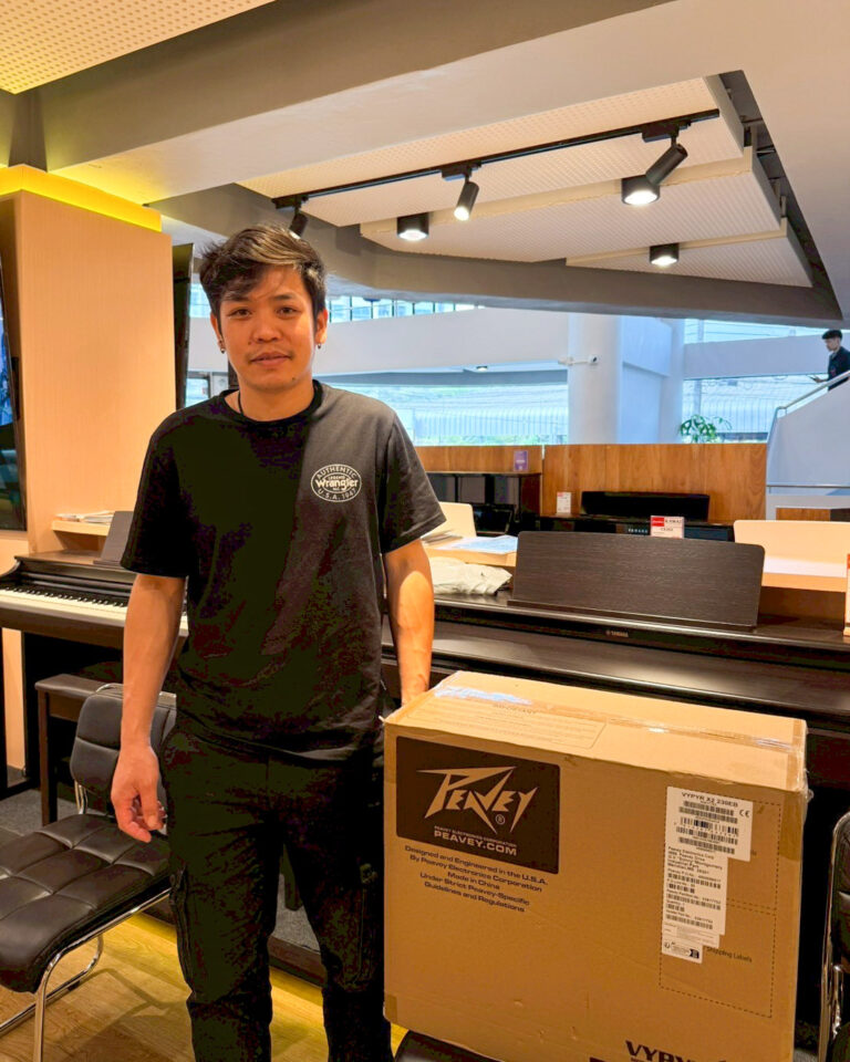 Man in a black t-shirt stands beside a large cardboard box in a music store, with pianos and chairs in the background.
