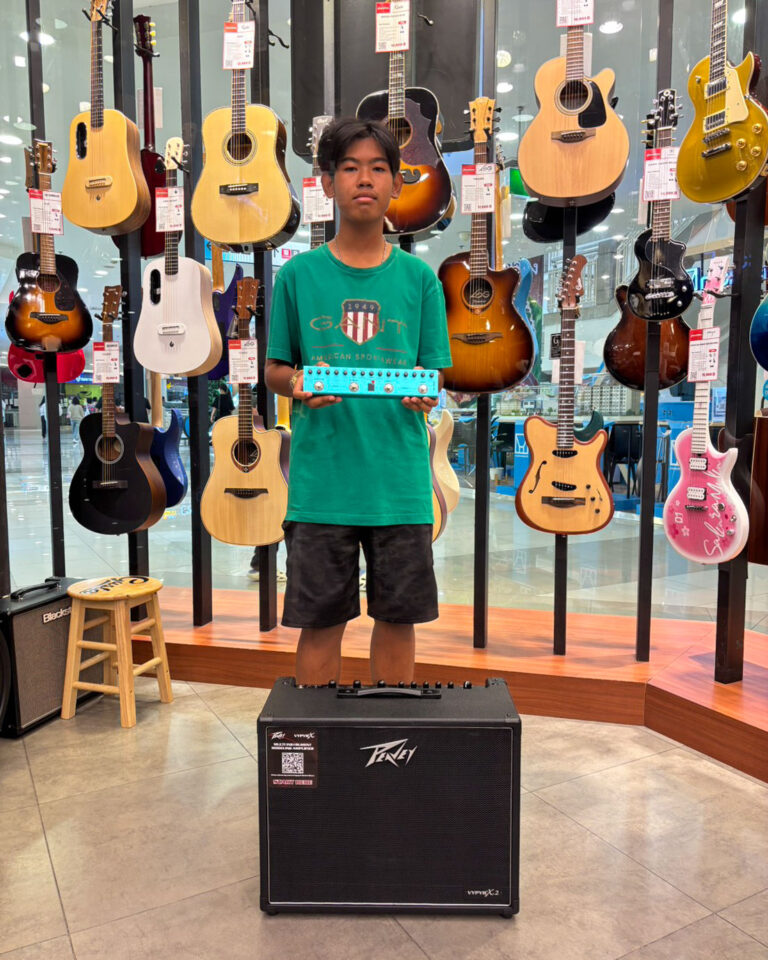 Boy in a guitar shop holding a blue multi-effect pedal, with acoustic guitars on display behind him.