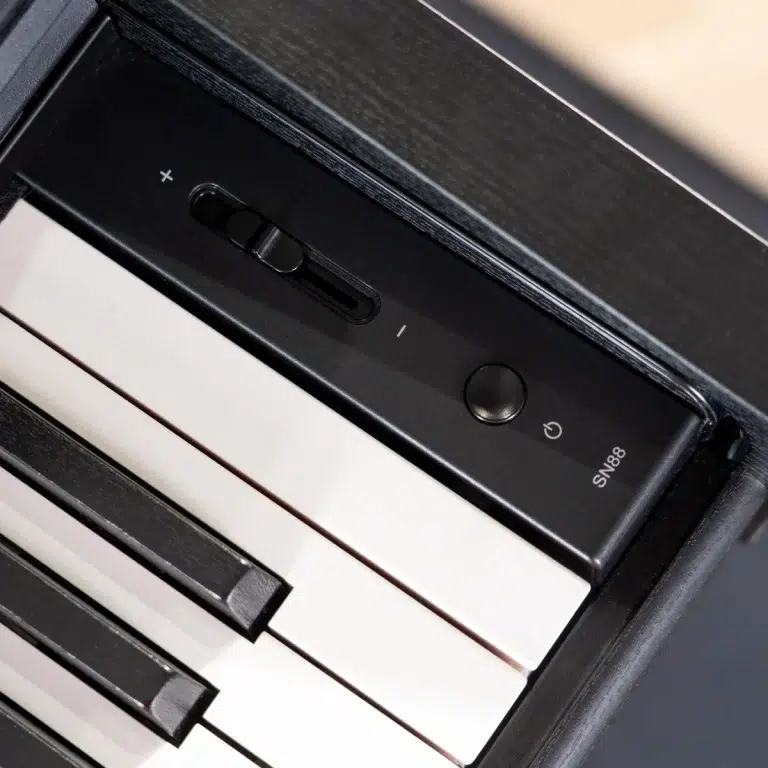Close-up of a digital piano keyboard showing a black control panel with a volume slider marked + and - and a circular power button, with white and black keys visible nearby. ขายราคาพิเศษ