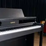 Black upright Samick piano with white keys, set against a dark curtain backdrop. ขายราคาพิเศษ