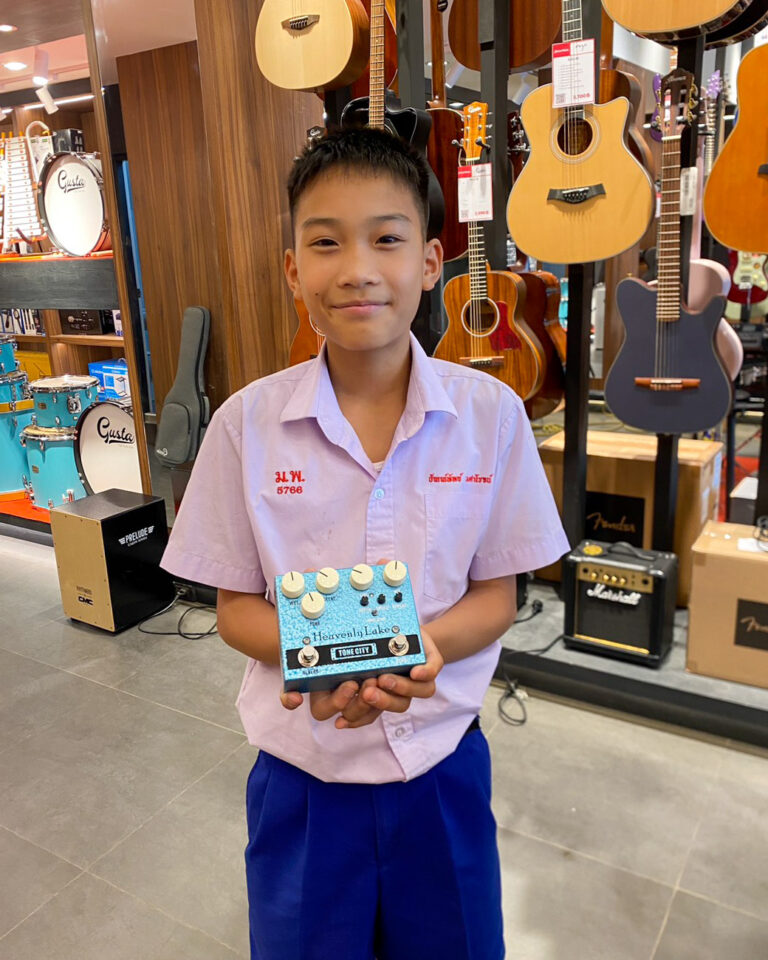 Smiling boy in a light purple shirt and blue shorts holds a blue guitar effects pedal in a guitar shop.