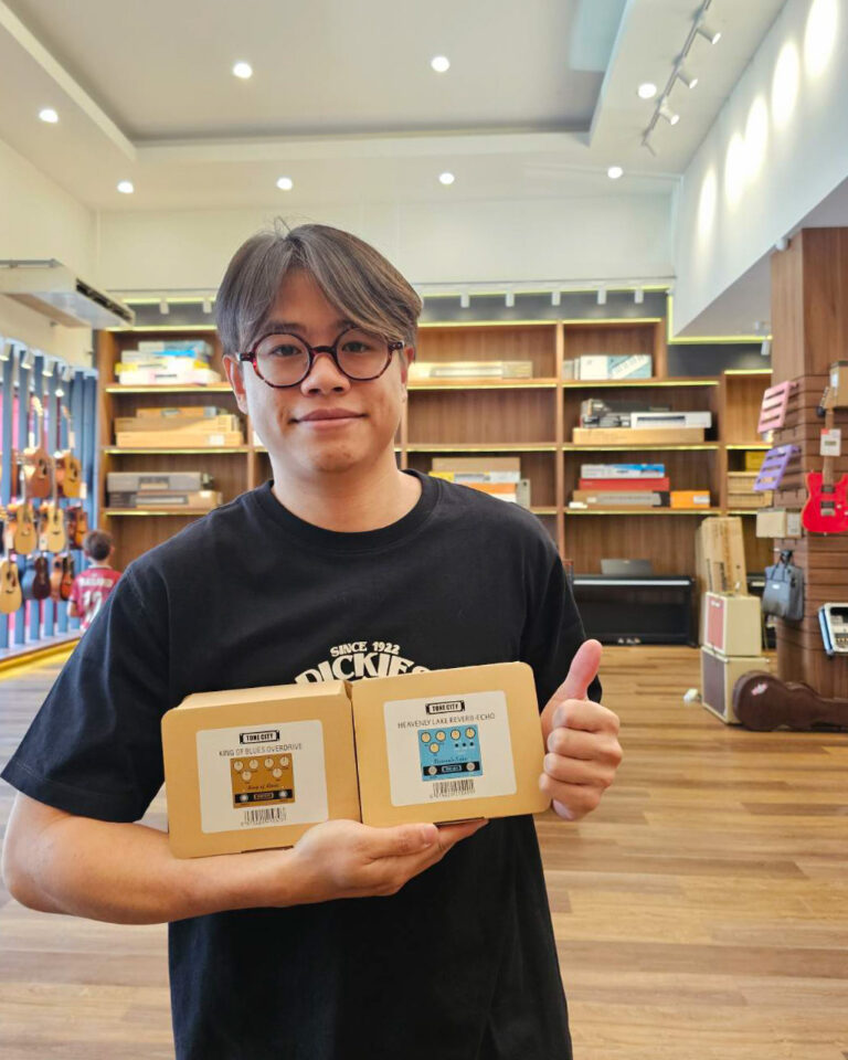 Young man with glasses in a music store, holding two small cardboard boxes with cassette-style labels and giving a thumbs up.