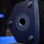 Close-up of a dark blue speaker cabinet with a circular port in the center, four screws around it, and rubber feet on a blue carpeted surface. ขายราคาพิเศษ