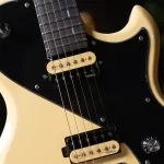 Close-up of a cream-colored electric guitar with a black pickguard, two cream Tesla pickups, and six strings visible across the fretboard nearby. ขายราคาพิเศษ