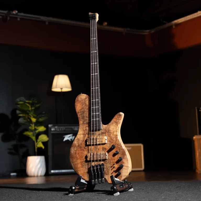 Wooden electric bass guitar on a stand in a dim studio, with a lamp and amp in the background. ขายราคาพิเศษ