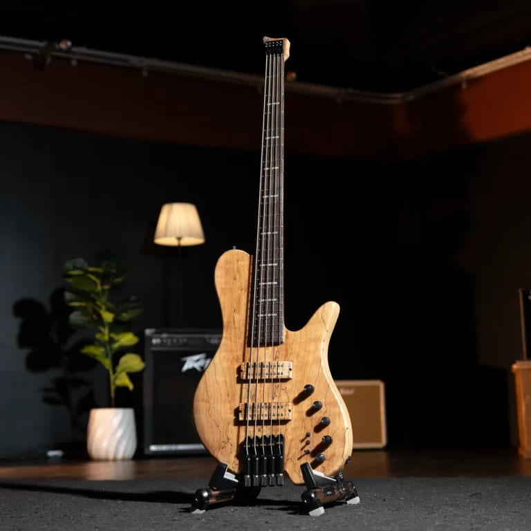 Natural wood electric bass guitar on a stand in a dim studio with amps and a plant in the background. ขายราคาพิเศษ