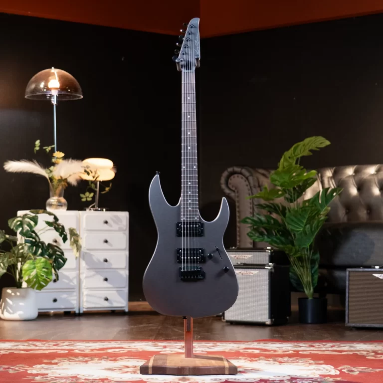 Gray electric guitar on a stand in a cozy studio with plants, a white dresser, and guitar amplifiers in the background. ขายราคาพิเศษ