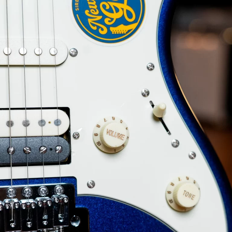 Close-up of a blue electric guitar body with a white pickguard, cream volume and tone knobs, pickups, and chrome hardware; a round blue-yellow sticker sits on the pickguard. ขายราคาพิเศษ
