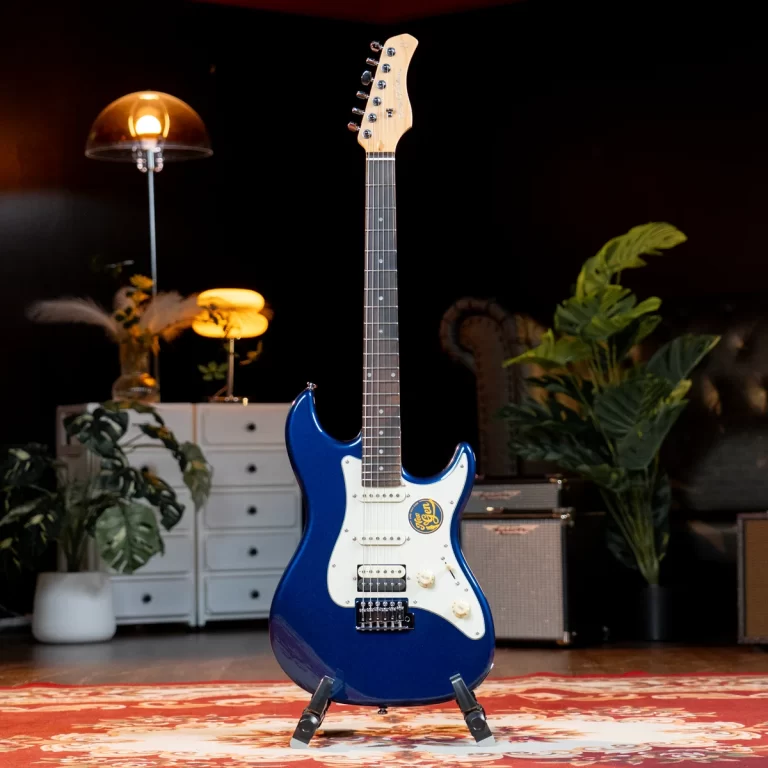 Blue electric guitar on a stand in a cozy studio setup with plants, a rug, and amps in the background. ขายราคาพิเศษ