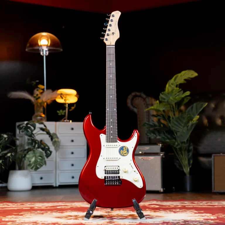 Red electric guitar on a stand in a cozy home studio with plants, a lamp, and amps in the background. ขายราคาพิเศษ