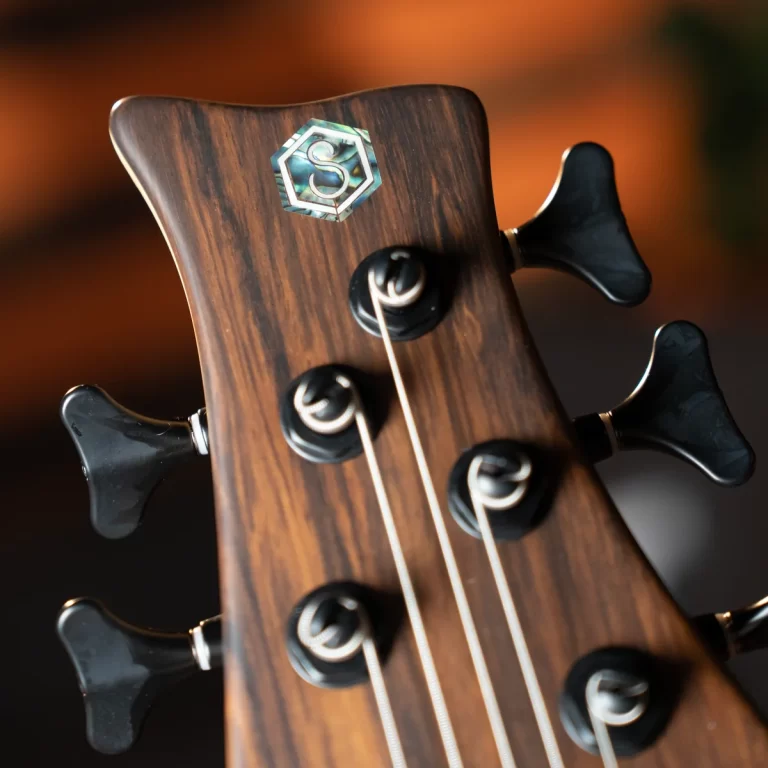 Close-up of a wooden guitar headstock with tuning pegs and strings; a hexagonal badge with an S logo. ขายราคาพิเศษ