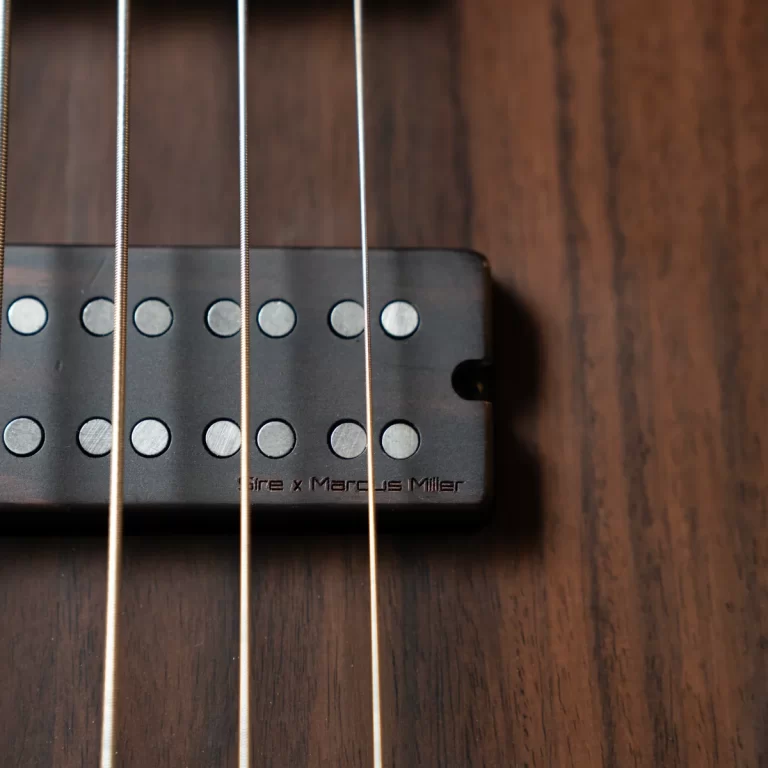 Close-up of a bass guitar pickup with metal pole pieces and four strings, labeled 'Sire x Marcus Miller' on a dark pickup plate over a wooden body surface. ขายราคาพิเศษ