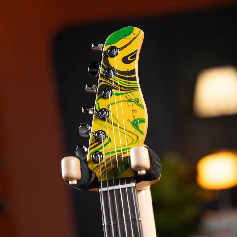 Close-up of a yellow and green marbled guitar headstock with tuning pegs, held by gloved hands in a dim room. ขายราคาพิเศษ