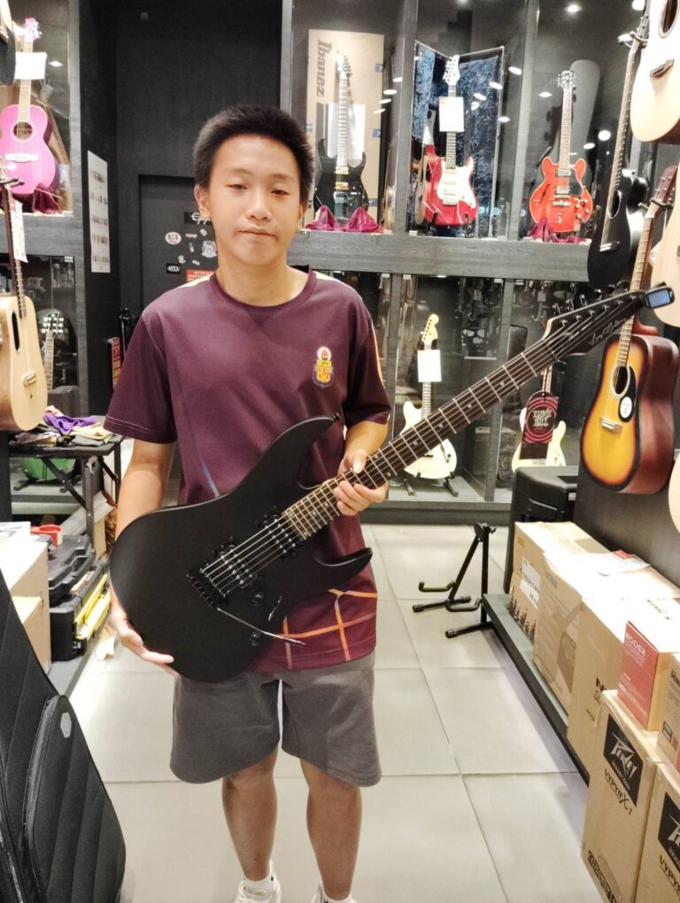 Young man in a music store holding a black electric guitar behind his body, looking at the camera.