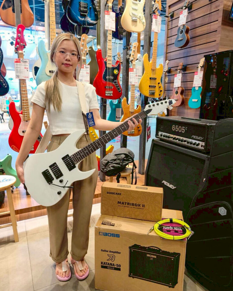 Person holding a white electric guitar in a guitar shop with colorful guitars on wall behind them and gear boxes nearby.