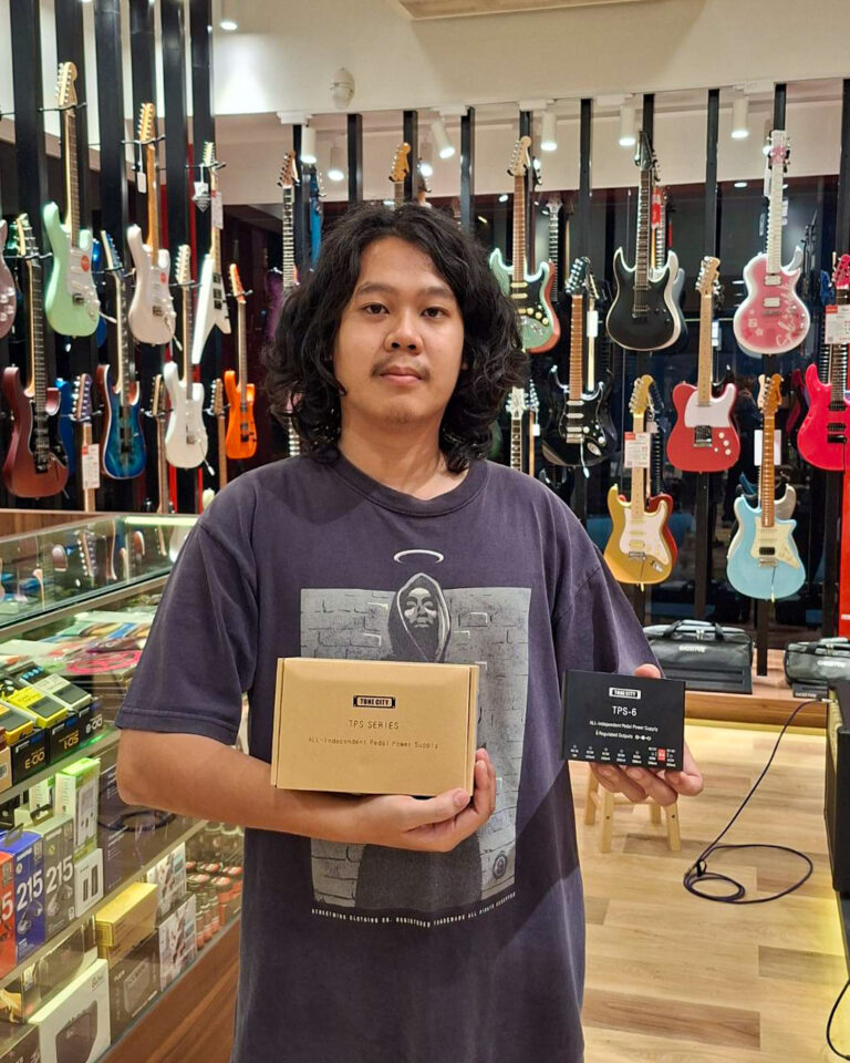 Man with long dark hair in a guitar store holds a tan TPS Series box and a smaller black box, guitars hanging in the background.