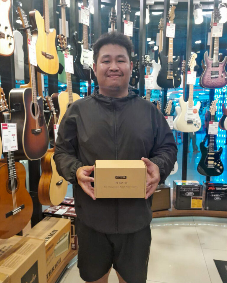 Smiling man in a black jacket stands in a guitar shop, holding a cardboard box labeled 'TPS SERIES'.