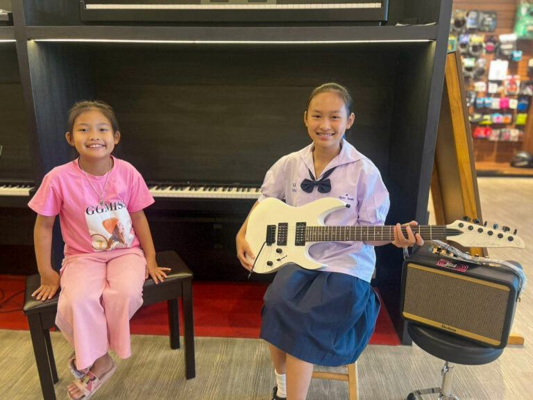 Two young girls in a music shop: one sits on a stool by a piano wearing pink clothing, the other sits on a chair holding a white electric guitar with an amplifier nearby and smiling at the camera.