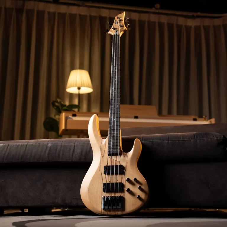 Wooden bass guitar leaning against a sofa in a cozy living room with a lamp and keyboard in the background. ขายราคาพิเศษ