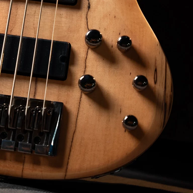 Close-up of a natural wood electric bass guitar body showing four chrome knobs and a bridge with strings. ขายราคาพิเศษ