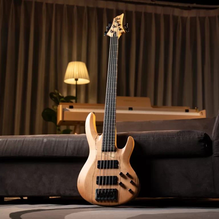 Natural-finish six-string bass guitar leaning against a dark sofa with a softly lit lamp and a keyboard in the background. ขายราคาพิเศษ