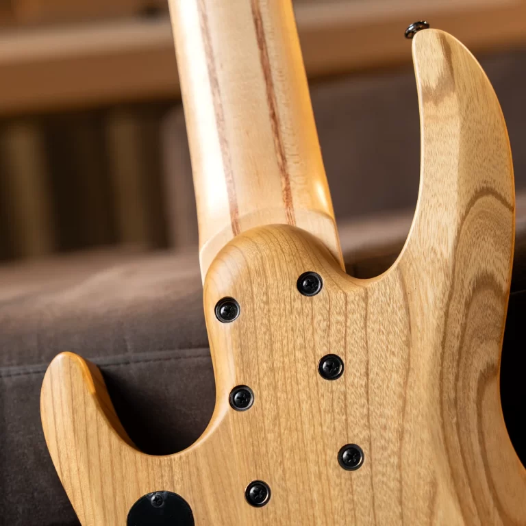 Close-up of a natural wood electric bass guitar body with a neck joint and black screws visible in the cavity area on a dark sofa surface. ขายราคาพิเศษ