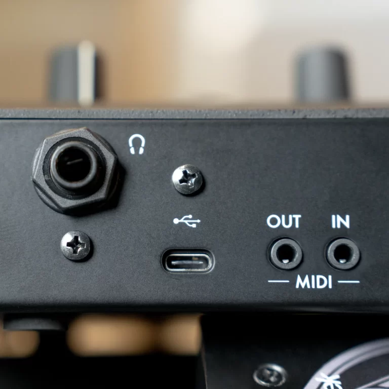 Close-up of a black audio interface front panel showing a large 1/4-inch jack, USB-C port, and MIDI OUT/IN sockets labeled OUT and IN. Two small screws secure the panel. ขายราคาพิเศษ