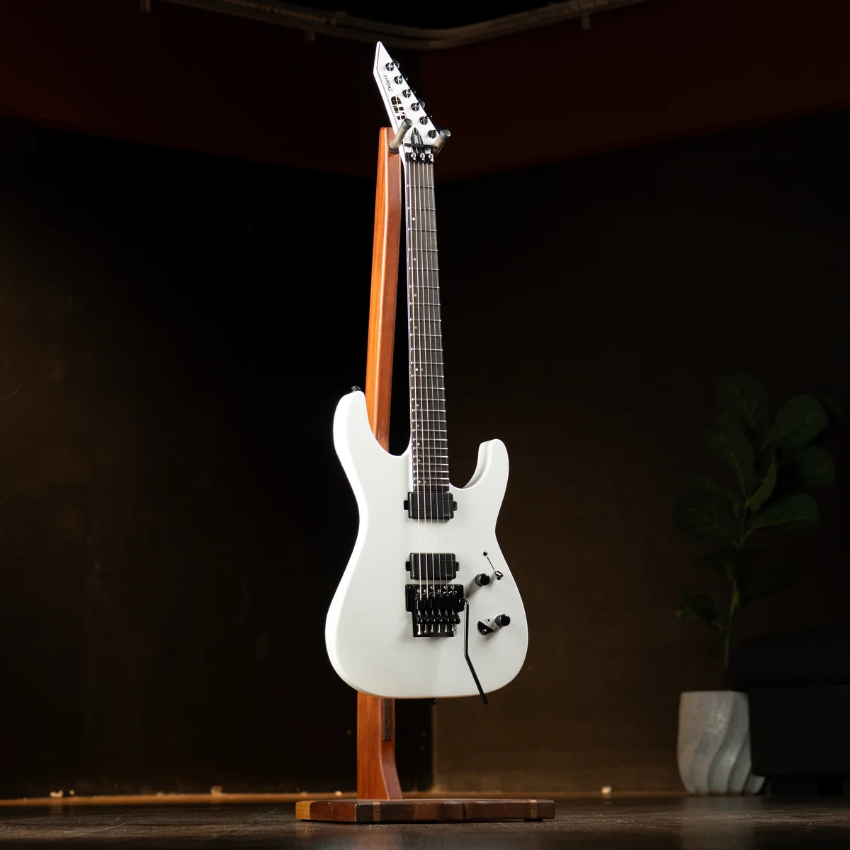 White electric guitar on a wooden stand in a dark studio with a plant in the background.