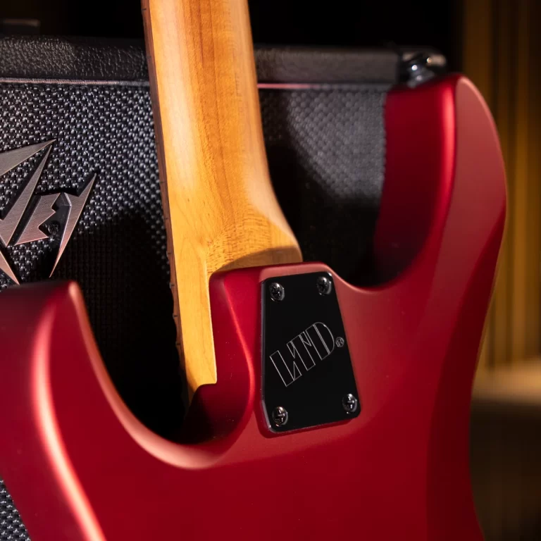 Close-up of a red electric guitar body with a natural wood neck resting against a textured black amplifier; metal plate with screws visible on the back of the guitar. ขายราคาพิเศษ