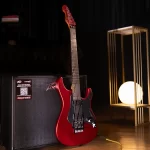 Red electric guitar leaning against a black amplifier on a wooden floor, with a warm lamp in a modern stage setup behind it. ลดราคาพิเศษ