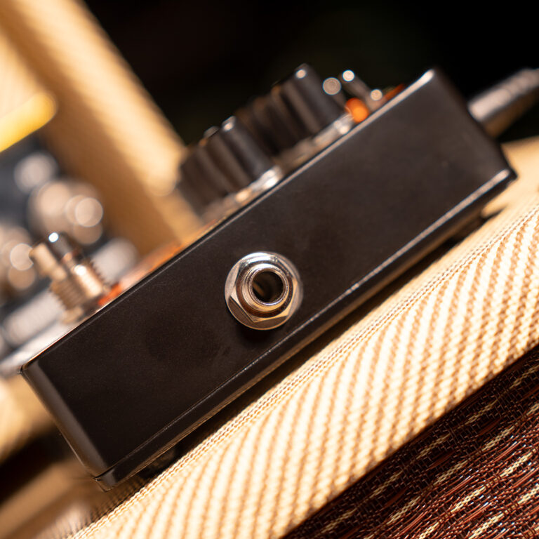 Close-up of a black guitar effects pedal on a striped guitar strap, focusing on a 1/4 inch input jack in the center of the panel. ขายราคาพิเศษ