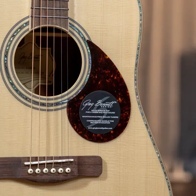 Close-up of an acoustic guitar body showing the sound hole, six strings, and a tortoiseshell pickguard with a Greg Bennett Design sticker inside the guard. ขายราคาพิเศษ