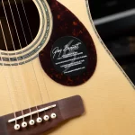 Close-up of an acoustic guitar body with the soundhole, strings, and a black circular sticker that reads 'Greg Bennett Design' near the pickguard. ขายราคาพิเศษ