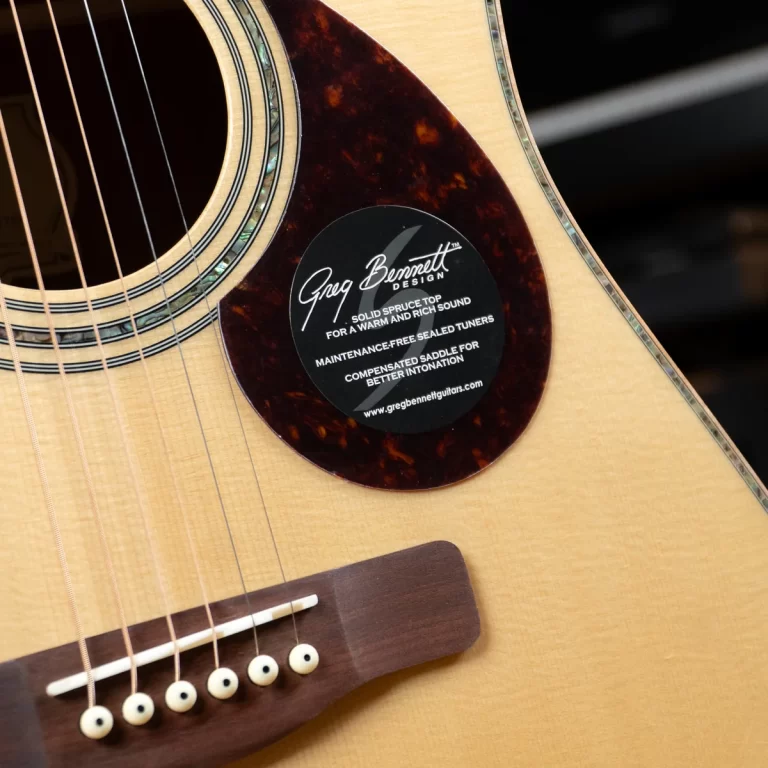 Close-up of an acoustic guitar body with the soundhole, strings, and a black circular sticker that reads 'Greg Bennett Design' near the pickguard. ขายราคาพิเศษ