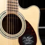 Close-up of an acoustic guitar body showing the soundhole, rosette, and light wood top with a tortoiseshell pickguard and decorative inlays around the edge. A sticker from Greg Bennett Design is visible on the lower right. ขายราคาพิเศษ