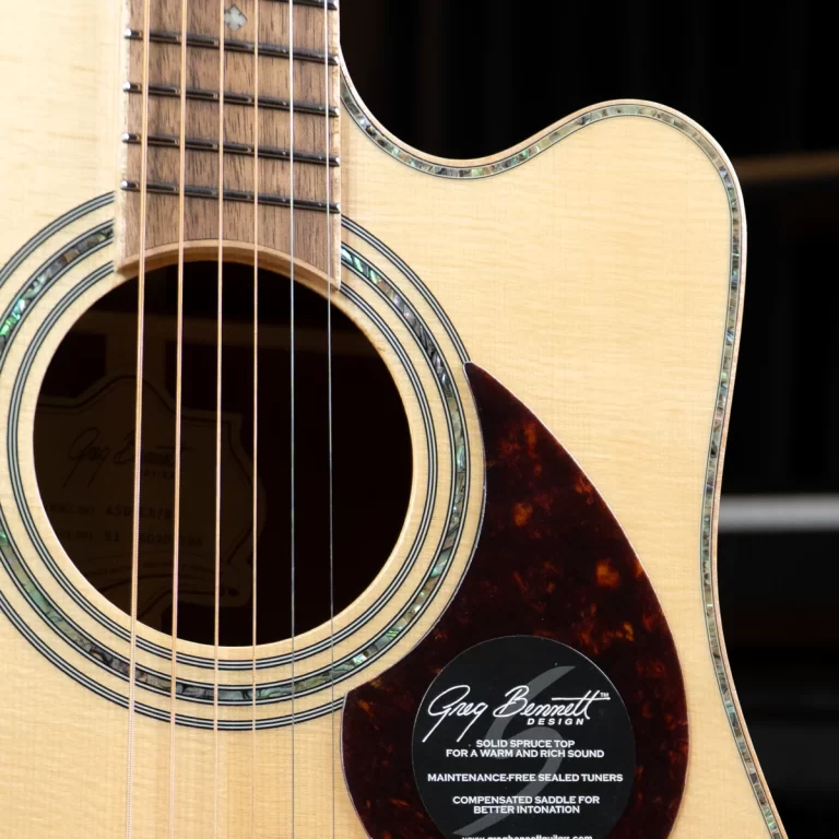 Close-up of an acoustic guitar body showing the soundhole, rosette, and light wood top with a tortoiseshell pickguard and decorative inlays around the edge. A sticker from Greg Bennett Design is visible on the lower right. ขายราคาพิเศษ