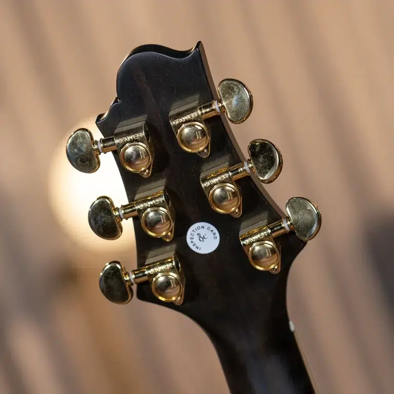 Close-up of a black guitar headstock with gold tuning pegs arranged along both sides of the neck, a small circular inspection sticker visible on the back. ขายราคาพิเศษ
