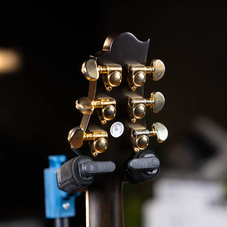 Close-up of a guitar headstock's back with gold tuning pegs and a small inspection sticker, blurred dark background ขายราคาพิเศษ