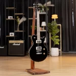 Black electric guitar on a wooden stand in a studio/showroom with amps, shelves, and potted plants in the background. ขายราคาพิเศษ