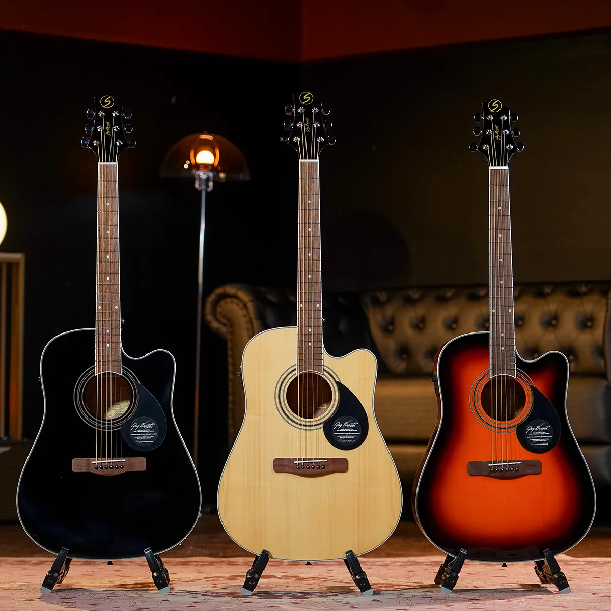 Three acoustic guitars on stands in a showroom, finished in black, natural, and sunburst against a dark backdrop, ready to play.