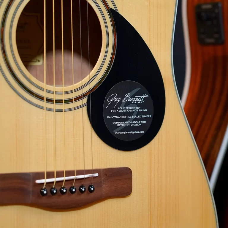 Close-up of an acoustic guitar showing the soundhole, strings, and a black circular sticker on the pickguard that reads 'Greg Bennett Design'. ขายราคาพิเศษ